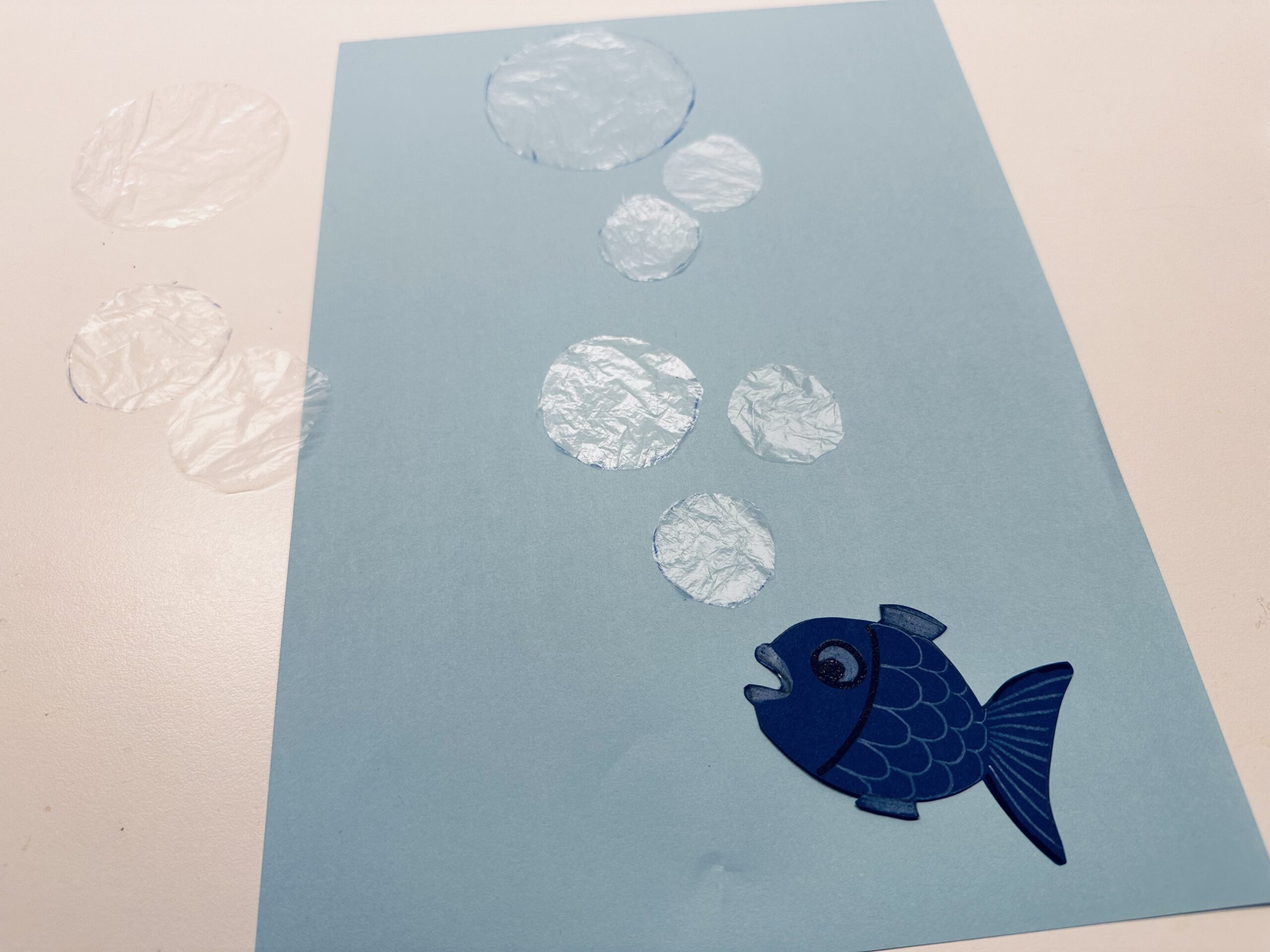 Bild zum Schritt 11 für das Bastel- und DIY-Abenteuer für Kinder: 'Legt den fertigen Fisch auf euer blaues Papier und ordnet...'